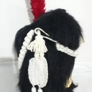 Puede incluir: Un sombrero militar negro con una pluma roja, ribete de cuerda blanca y borlas blancas. El sombrero tiene una textura similar a la piel y detalles dorados. Tocado histórico.