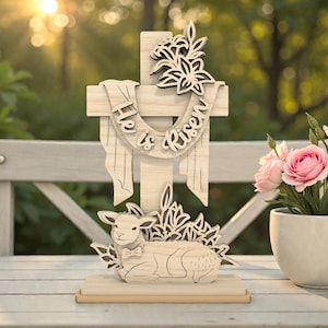 Puede incluir: Decoración de Pascua de madera con una cruz con la inscripción "Él ha resucitado" y un cordero. La cruz está adornada con detalles florales. Un jarrón con rosas rosas está cerca. La escena está ambientada al aire libre con luz natural.