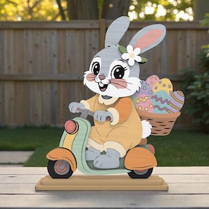 Può includere: Figurina in legno di un coniglietto pasquale su uno scooter, con un cestino di uova decorate. Il coniglio è grigio con un fiore, indossa un abito arancione. Lo scooter è verde menta e arancione, su una base di legno. Perfetto per le decorazioni pasquali.