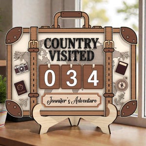 Può includere: Contatore da viaggio in legno a forma di valigia, con la scritta "COUNTRY VISITED" e il numero "034". Presenta illustrazioni a tema viaggio e il nome "Jennifer's Adventure". Il design è nei toni del marrone e crema.
