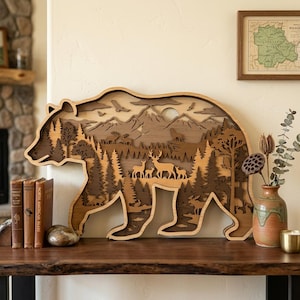 May include: A layered wooden bear-shaped wall art piece. The artwork features a detailed forest scene with trees, mountains, deer, and birds. The bear is in shades of brown and tan, creating a natural, rustic aesthetic. The piece is displayed on a wooden surface.