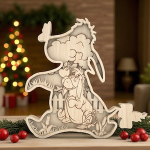 May include: Wooden layered decoration of Eeyore from Winnie the Pooh. The light-coloured wood carving shows Eeyore sitting, with a fence, clouds, and grass details. The decoration is placed on a wooden surface with red berries and greenery.