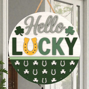 May include: Round wooden door sign with the words "Hello Lucky" in gray and green. The sign features a horseshoe and shamrock design, with a green and white color scheme. A rope is attached for hanging.