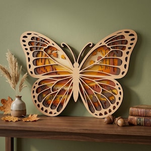 Puede incluir: Una gran pieza de arte mural de mariposa de madera en capas. La mariposa presenta un diseño detallado con recortes que revelan un paisaje otoñal en capas en tonos naranja, amarillo y marrón. La obra de arte se exhibe sobre una superficie de madera.