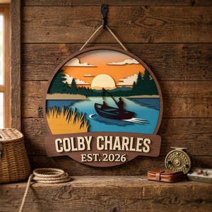 Könnte beinhalten: Rundes Holzschild mit einer Angelszene, das ein Boot auf einem blauen See vor einem Sonnenuntergang zeigt. Das Schild trägt die Aufschrift "COLBY CHARLES EST. 2026" in einem geschichteten Design und wird von einem Seil gehalten.