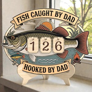 Pode incluir: Placa de madeira com design de peixe e os textos "FISH CAUGHT BY DAD" e "HOOKED BY DAD". A placa mostra o número 126. É suportada por um suporte de madeira e apresenta um peixe, água e uma fogueira.