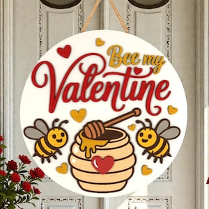 May include: A circular wooden sign with the words "Bee my Valentine" in red and gold. The sign features a honey pot, two bees, and heart accents. A rope is attached for hanging.
