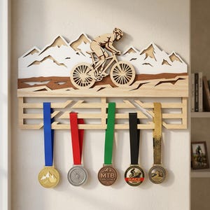 May include: Wooden medal display with a cyclist and mountain range design. Below, five medals with coloured ribbons hang from a wooden rack. The medals have gold, silver, and bronze tones. The rack is approximately 60 cm wide.
