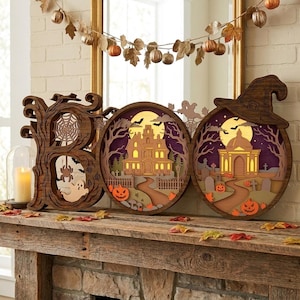 Op de afbeelding: Een decoratief houten bord met de tekst BOO, bestaande uit gedetailleerde, lasergesneden lagen. De B bevat een spinnenweb en een geest, terwijl de twee O's verlichte scènes tonen van een spookhuis, begraafplaats, pompoenen en een heksenhoed.