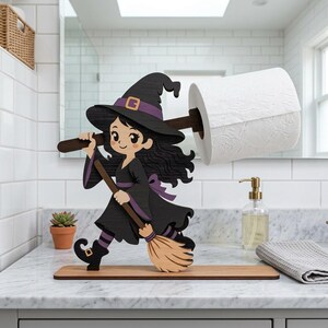 May include: A wooden witch-themed toilet paper holder. The black witch figure is wearing a purple hat and dress, holding a broom. The toilet paper roll is held by a wooden dowel. The holder is on a wooden base.