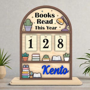 May include: A wooden desk organizer with the text "Books Read This Year" and the number "128". The organizer has a brown frame, a light wood background, and blue lettering that says "Kento". Decorative elements include a cactus, books, and a desk lamp.