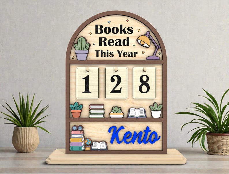 May include: A wooden desk organizer with the text "Books Read This Year" and the number "128". The organizer has a brown frame, a light wood background, and blue lettering that says "Kento". Decorative elements include a cactus, books, and a desk lamp.