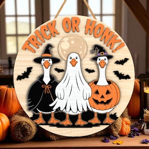 May include: Round wooden Halloween sign with the text "TRICK OR HONK!" in orange. The sign features three geese dressed in Halloween costumes: a witch, a ghost, and a jack-o'-lantern. Black bats and a full moon are also included in the design.