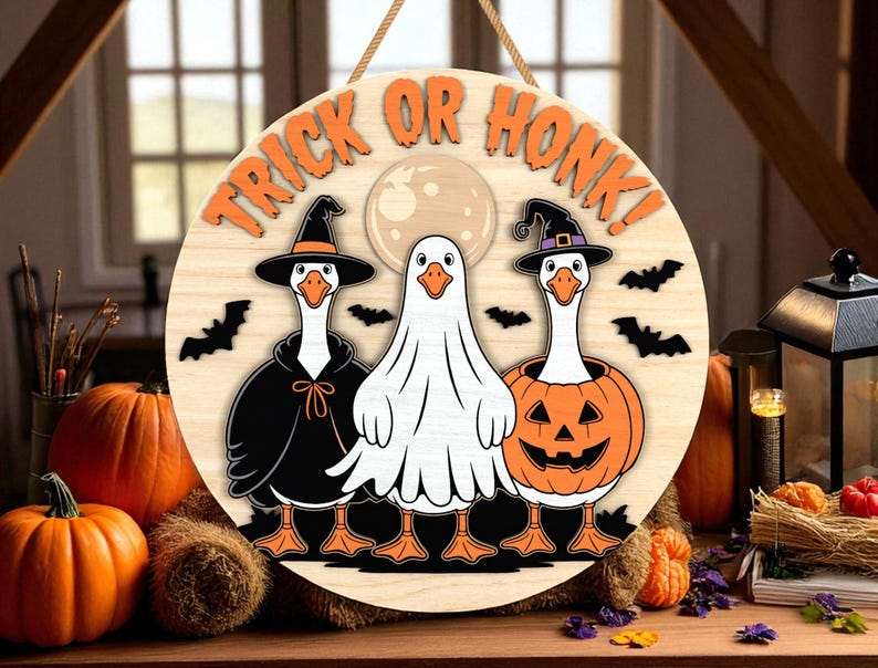 May include: Round wooden Halloween sign with the text "TRICK OR HONK!" in orange. The sign features three geese dressed in Halloween costumes: a witch, a ghost, and a jack-o'-lantern. Black bats and a full moon are also included in the design.