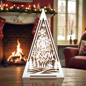 May include: A white, laser-cut Christmas tree decoration with the words "Let it Snow" and a winter scene. The tree features snowflakes, stars, trees, mountains, and a small house. The decoration is placed on a wooden surface.