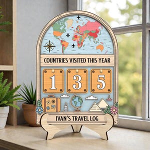 May include: Wooden travel log with a world map, airplanes, and the text "COUNTRIES VISITED THIS YEAR." It displays "135" in a counter format. The base reads "IVAN'S TRAVEL LOG." Decorative elements include a suitcase, globe, and landmarks.