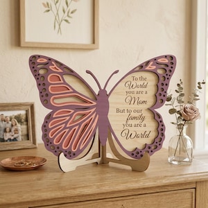 Op de afbeelding: Een houten decoratief vlindervormig ornament met gelaagde vleugels in paarse en roze tinten. Het middenstuk heeft de tekst "To the World you are a Mom, But to our family you are a World". Het staat op een houten standaard.