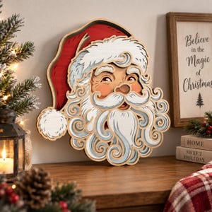 May include: A layered wooden wall decoration featuring the face of Santa Claus with a white beard, moustache, and eyebrows, wearing a red hat with a white pom-pom. The design uses stacked wood cutouts to create depth and dimension in the facial features.
