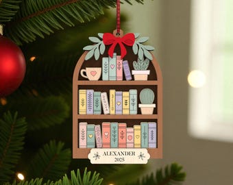 May include: Wooden Christmas tree ornament in the shape of a bookshelf, with a red bow, books, a teacup, and a cactus. The ornament is personalised with the name "ALEXANDER" and the year "2025". The ornament is hanging on a Christmas tree.