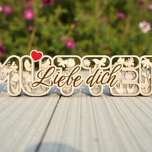 May include: Wooden sign with the word "MUTTER" and the phrase "Liebe dich" in a decorative font. The letters are filled with floral and butterfly cutouts, with a red heart above the "U".