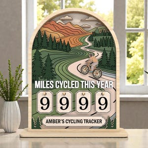 May include: Wooden cycling tracker with a mountain and road design. The text "MILES CYCLED THIS YEAR" is above the counter, which displays "9999". The bottom of the tracker reads "AMBER'S CYCLING TRACKER".