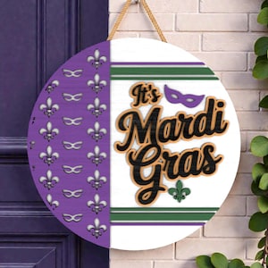 May include: A round wooden sign with a Mardi Gras theme. The left side is purple with fleur-de-lis and mask cutouts. The right side is white with green and purple stripes and the words "It's Mardi Gras" in black and gold.