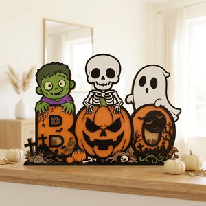 May include: A wooden Halloween decoration featuring the word BOO, where the letter B is topped by a green zombie, the letter O is a carved orange jack-o'-lantern with a skeleton on top, and the second O features a white ghost and a moon.