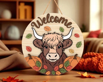 May include: Round wooden welcome sign featuring a Highland cow with long white horns, surrounded by fall leaves and pumpkins. The word "Welcome" is in brown letters at the top. A jute rope is attached for hanging.