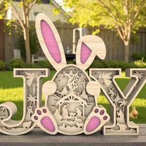 Puede incluir: Decoración de Pascua de madera que deletrea "JOY" con un diseño de oreja de conejo y una escena de la Natividad dentro del conejo. Las letras son de color marrón claro con acentos rosados. La decoración está colocada sobre una superficie de madera.