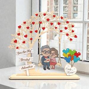 May include: A wooden decorative piece featuring a tree with red heart-shaped leaves and a swing with an elderly couple. The sign reads "You Are My Greatest Adventure". Colorful balloons and a heart-shaped base complete the design.