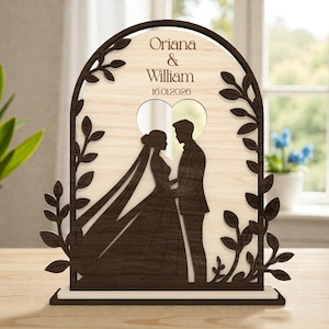 Pode incluir: Placa de casamento em madeira com silhueta de noivos dentro de um recorte em forma de coração. A placa inclui os nomes "Oriana & William" e a data "16.01.2026", num design de arco decorativo com detalhes de folhas.