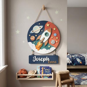 May include: A wooden space-themed wall hanging with a rocket ship, planets, and stars. The sign has a blue background with the name "Joseph" and "EVANS" in white lettering. The sign is hanging on a wall.