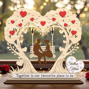 May include: A wooden heart-shaped tree with a swing, featuring a couple silhouette. Red heart accents adorn the tree. The base reads "Together is our favourite place to be." A heart-shaped sign displays names. Roses flank the display.