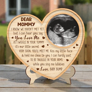 May include: A heart-shaped wooden plaque with a sonogram image, and the text "DEAR MOMMY" and a loving message. The plaque is supported by a wooden stand. The text includes the phrase "You Love Me" and "Love, BABY BUMP".