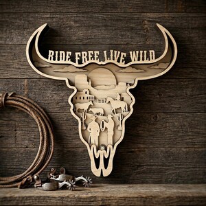 Puede incluir: Arte de pared de madera con forma de calavera de toro con las palabras "RIDE FREE LIVE WILD". La obra de arte presenta un diseño en capas con una escena del oeste de vaqueros, caballos y ganado. Una cuerda y espuelas están en primer plano.