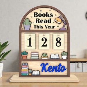May include: Wooden bookshelf-style desk organizer with the words "Books Read This Year" and the number "128". The name "Kento" is displayed in blue. Decorative elements include a cactus, a lamp, and small books.