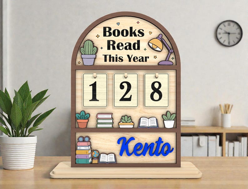May include: Wooden bookshelf-style desk organizer with the words "Books Read This Year" and the number "128". The name "Kento" is displayed in blue. Decorative elements include a cactus, a lamp, and small books.