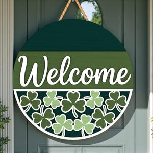 May include: A round, green wooden welcome sign with a rope hanger. The sign has a dark green top, a lighter green middle section with the word "Welcome" in white script, and a bottom section with shamrock cutouts.