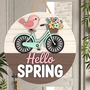 Puede incluir: Un letrero redondo de madera con temática primaveral. Presenta una bicicleta azul claro con una cesta de flores, un pájaro rosa y las palabras "Hello SPRING" en rosa y blanco. El letrero está colgado de una cuerda.