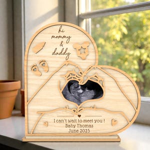 Könnte beinhalten: Holz-Fotorahmen in Herzform mit Laserschnittdetails. Der Rahmen enthält den Text "hi mommy & daddy" und "I can't wait to meet you! Baby Thomas June 2025". Der Rahmen enthält ein schwarz-weißes Ultraschallbild.