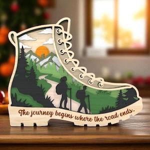 Può includere: Un'opera d'arte in legno a strati a forma di scarpa da trekking. L'interno raffigura una scena di montagna con escursionisti, alberi e un tramonto. Il testo "The journey begins where the road ends" è scolpito alla base.