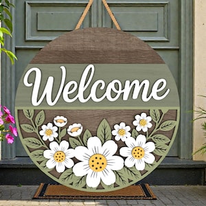 May include: Circular wooden welcome sign with a floral design. The sign has the word "Welcome" in white script, with white daisies and green leaves. It is suspended by a rope.