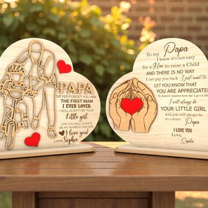 Op de afbeelding: Twee houten hartvormige plaquettes met lasergesneden ontwerpen en tekst. Eén toont een vader en kind op een fiets, de andere handen die een hart vasthouden. Beide hebben het woord "Papa" en liefdevolle berichten.
