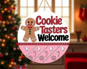 May include: A round, white wooden sign with the text "Cookie Tasters Welcome" in red and black. A gingerbread man is on the left. The bottom section is pink with gingerbread man, candy cane, and snowflake cutouts. A gold string is attached for hanging.
