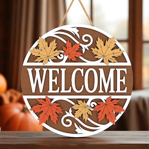 May include: A round wooden welcome sign with a brown background and white cut-out details. The word "WELCOME" is prominently displayed. Decorative maple leaves in orange and yellow are arranged around the sign, with a natural rope for hanging.