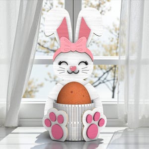 May include: A white wooden Easter bunny decoration with pink accents, holding a brown egg. The bunny has pink ears, a pink bow, and pink paw-shaped feet. The decoration is placed in front of a window with white curtains.