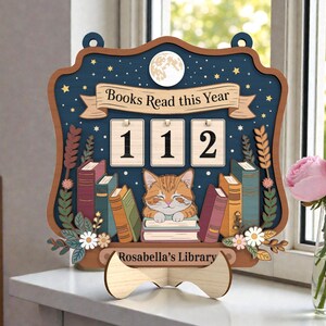 May include: Wooden book counter with a navy blue background, a full moon, and stars. The counter reads "Books Read this Year" and displays "112". A sleeping orange tabby cat rests on a stack of books. The counter is labeled "Rosabella's Library".