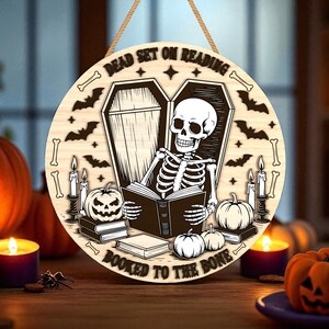 May include: Round wooden Halloween sign with a skeleton reading a book in a coffin, surrounded by bats, candles, and pumpkins. The sign reads "Dead Set on Reading" and "Booked to the Bone". Includes a jute rope for hanging.
