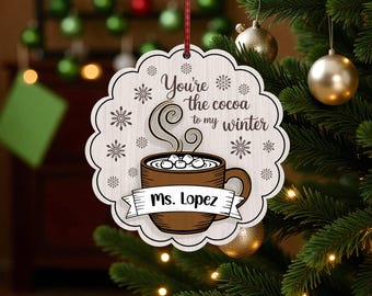 Op de afbeelding: Een houten kerstornament met een geschulpte rand, met een ontwerp van een warme chocolademok en de tekst "You're the cocoa to my winter". Het ornament bevat de naam "Ms. Lopez" op een banner. Sneeuwvlokken zijn ook inbegrepen.