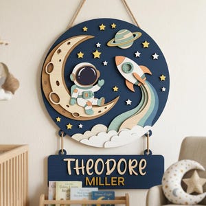 Puede incluir: Adorno de pared de madera azul marino con un diseño circular que representa un astronauta en una luna creciente, un cohete, estrellas y un planeta. Debajo, un letrero rectangular con el nombre "THEODORE MILLER". Ideal para una habitación infantil.